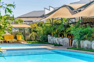 Piscina con sillas de descanso y sombrillas, rodeada de vegetación y un muro de piedra.