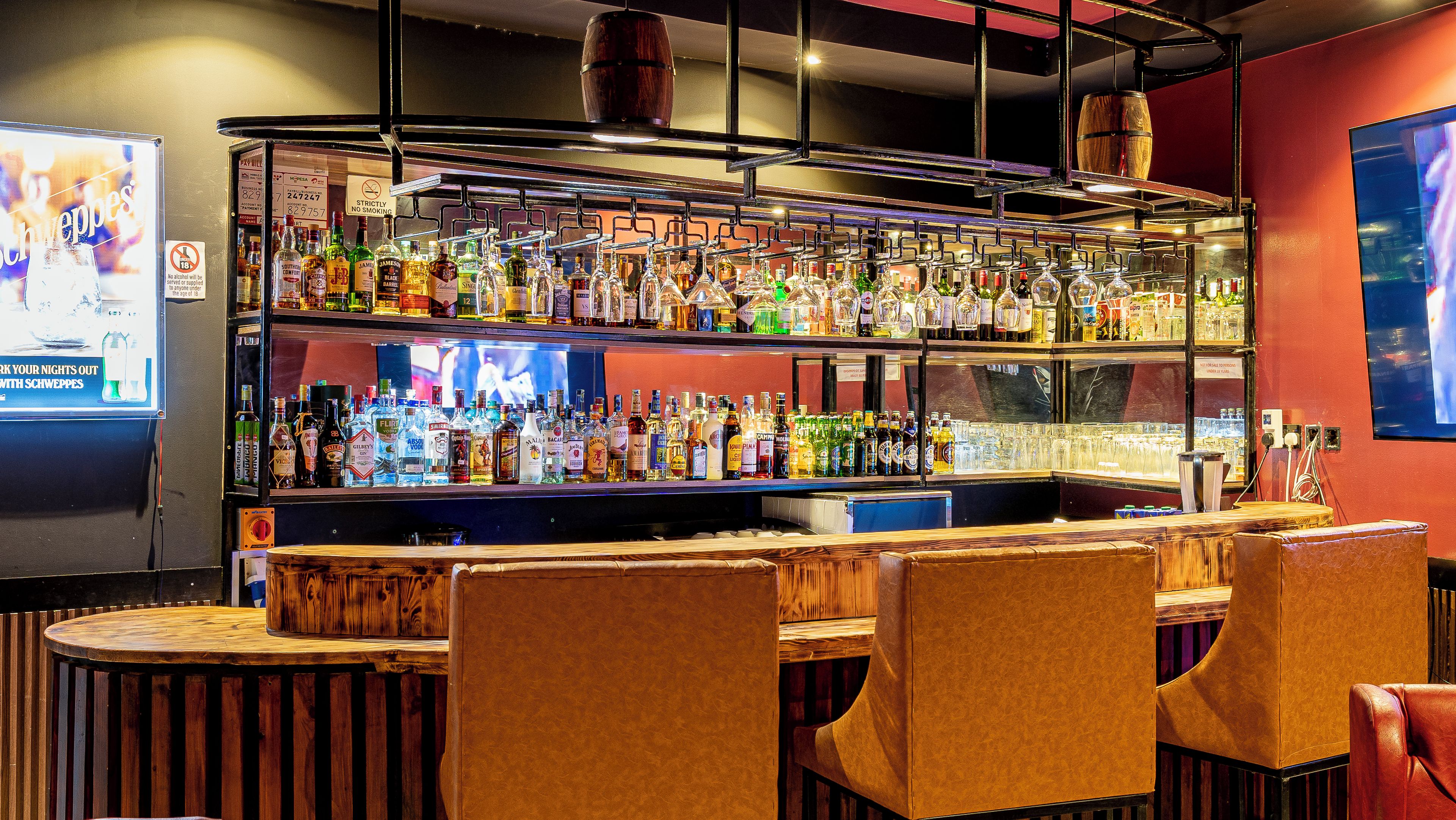 A bar area with multiple shelves of liquor bottles, a television screen, and leather bar stools.