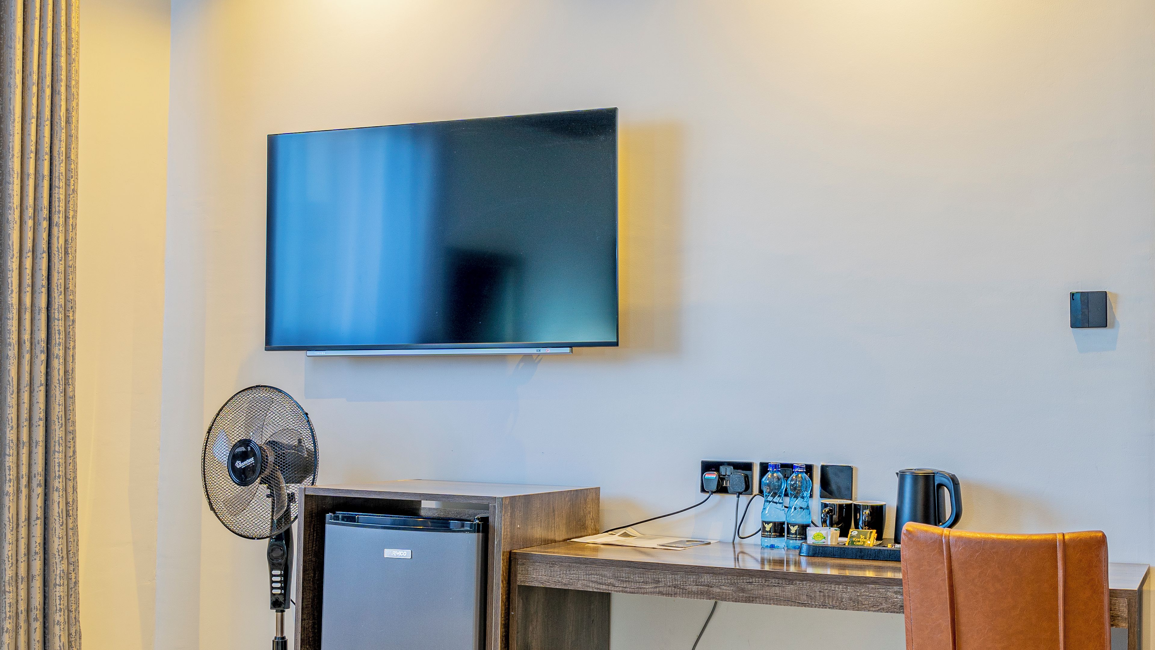 A hotel room with a flat-screen TV mounted on the wall, a small refrigerator, a desk with a chair, and a standing fan.