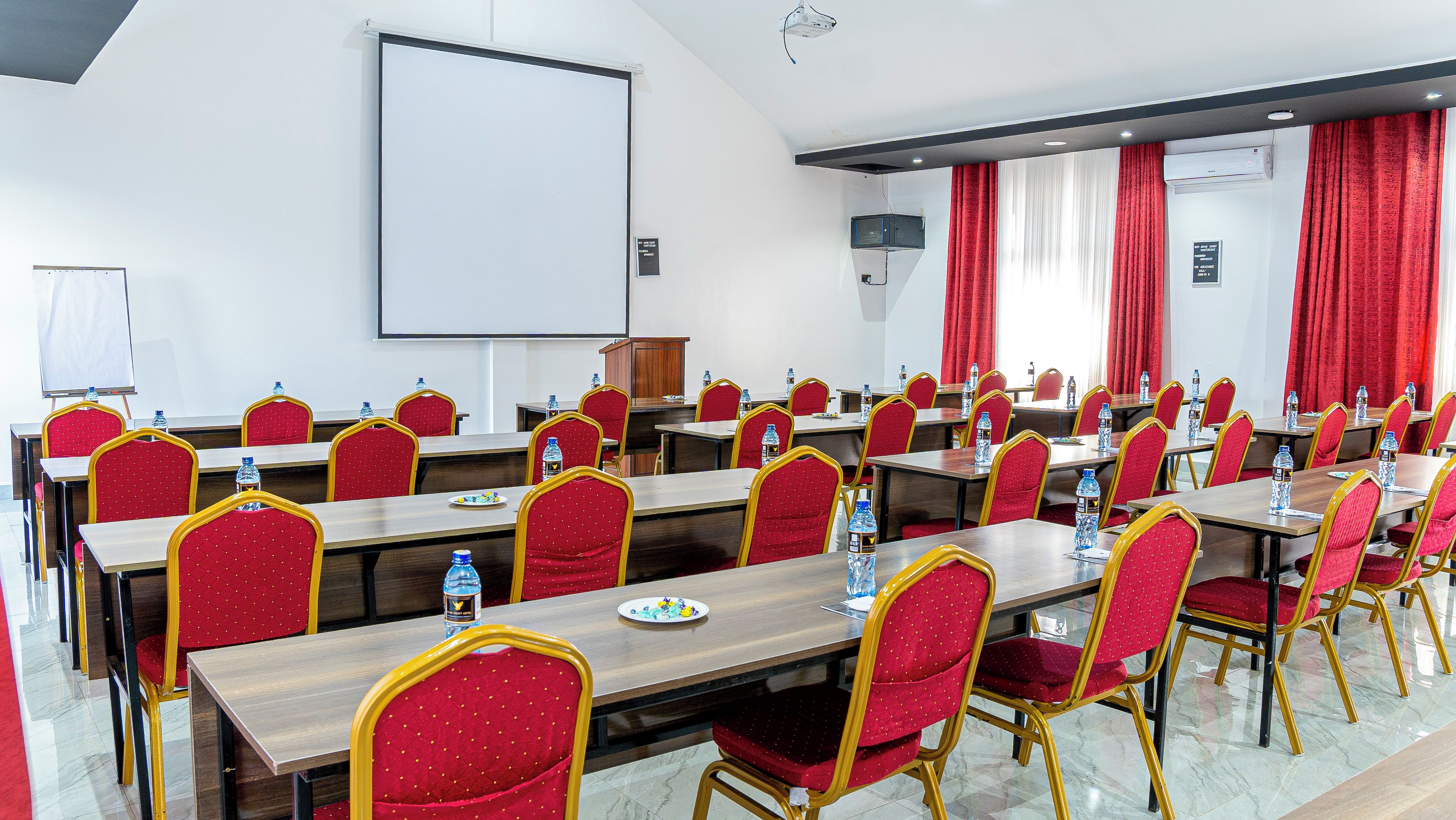 Un salón de conferencias con varias mesas y sillas, una pizarra y una pantalla de proyector.