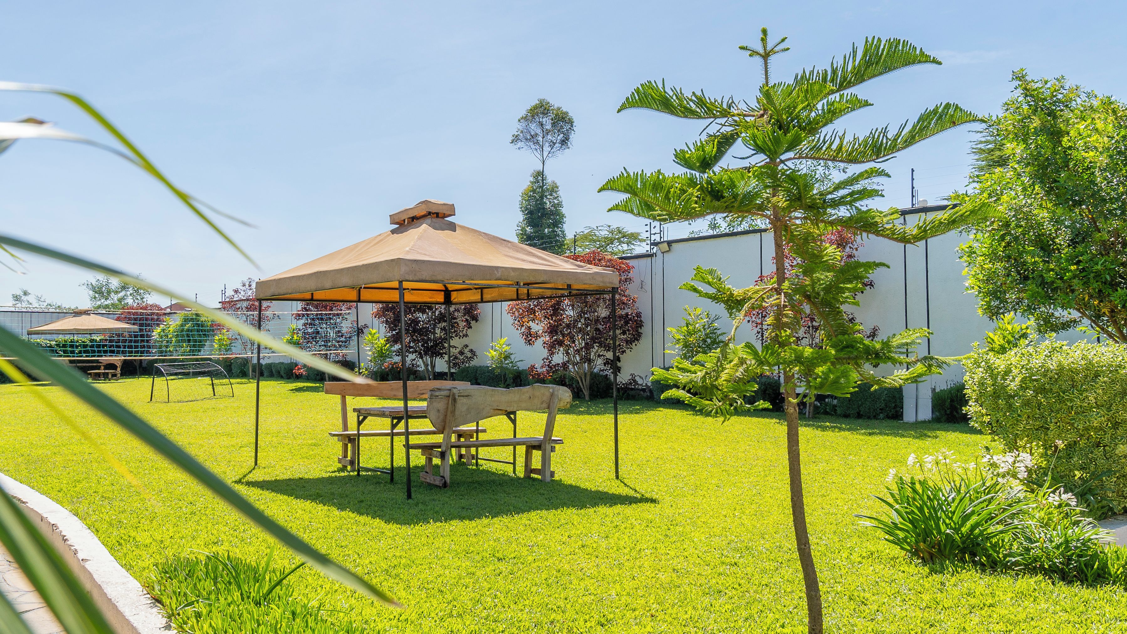 A well-maintained garden with a wooden table and chairs under a canopy, surrounded by lush greenery and a building in the background.