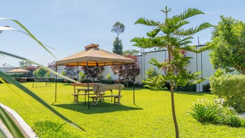 Un jardín bien cuidado con una mesa y sillas de madera bajo una pérgola, rodeado de exuberante vegetación y un edificio al fondo.