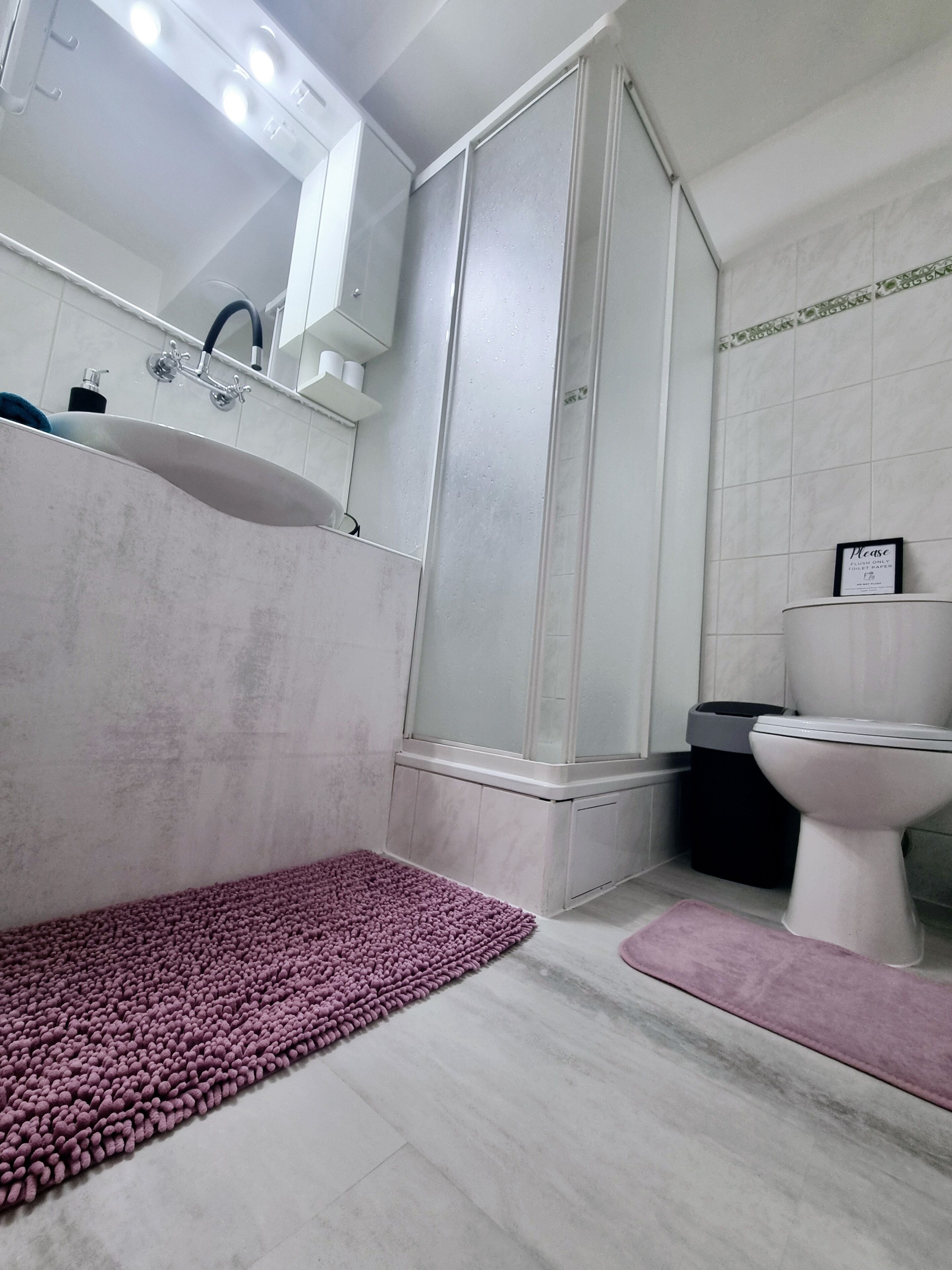 A bathroom with a sink, mirror, shower, toilet, and a purple rug.