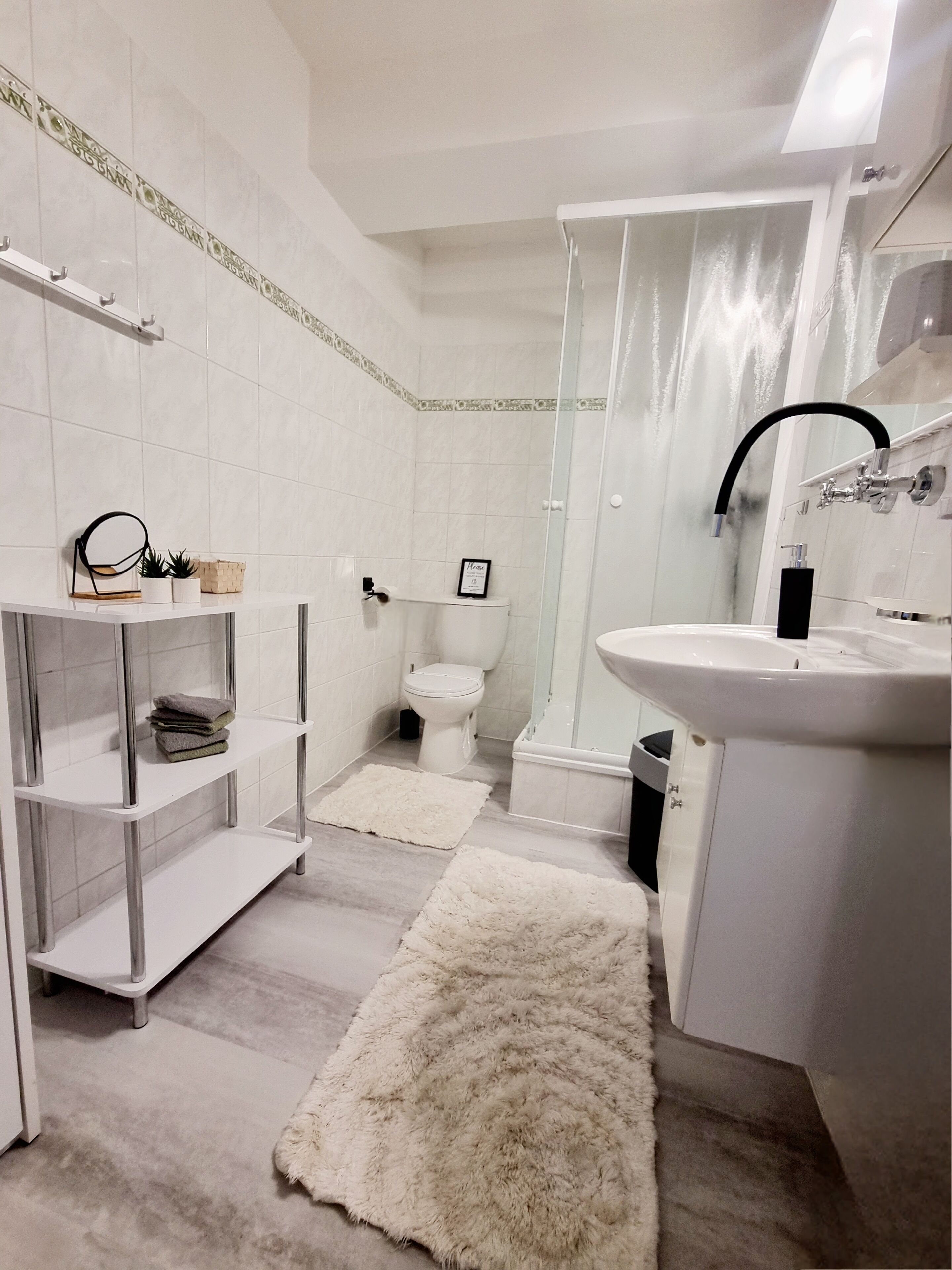 Un baño moderno con lavamanos, inodoro y ducha blancos. Hay estantes blancos con toallas y una alfombra mullida en el suelo.