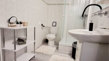 Un baño moderno con lavamanos, inodoro y ducha blancos. Hay estantes blancos con toallas y una alfombra mullida en el suelo.
