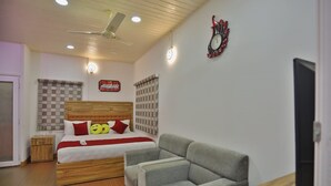 A bedroom with a bed, a grey sofa, a wooden nightstand, a TV, and a ceiling fan. - The Svarga Hill View Resort (Devikolam)