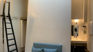 Une chambre d’hôtel moderne avec un lit, une échelle, une salle de bain avec un lavabo et un miroir, et une grande fenêtre.