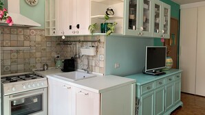 A kitchen with white cabinets, a stove, and a sink. There is a teal cabinet with a TV and a plant. - Bright & Pop Flat Isola & Central Station Hub (Milan)