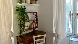 Une pièce avec un bureau en bois, une chaise blanche et un lit. On y trouve une étagère avec des plantes et des livres, ainsi qu’une fenêtre avec des rideaux.