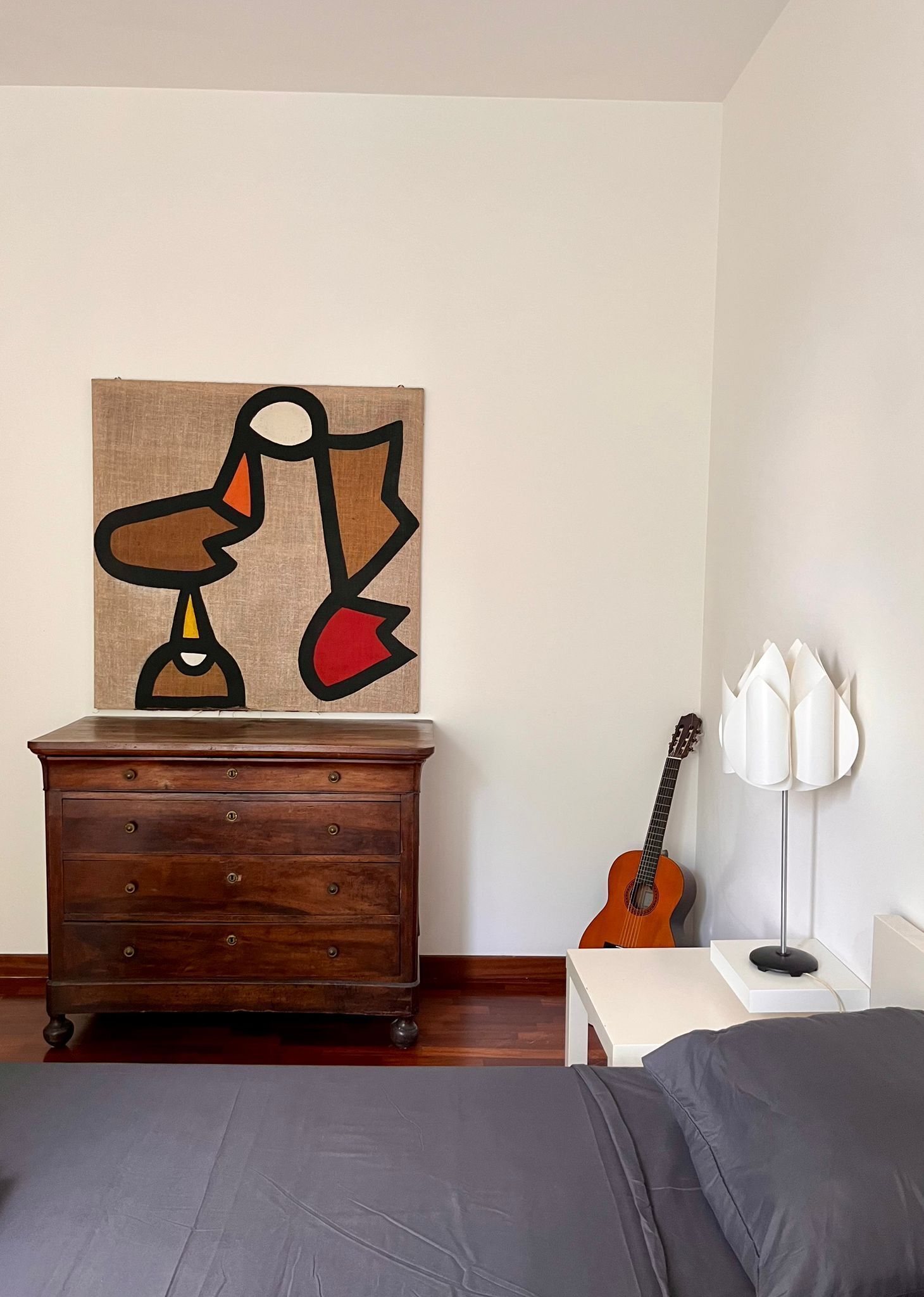 A bedroom with a wooden dresser, a guitar, a lamp, and a bed.