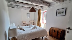 A bedroom with a wooden floor, a bed with white linens, a wooden chest, a hanging lamp, and a framed picture on the wall.
