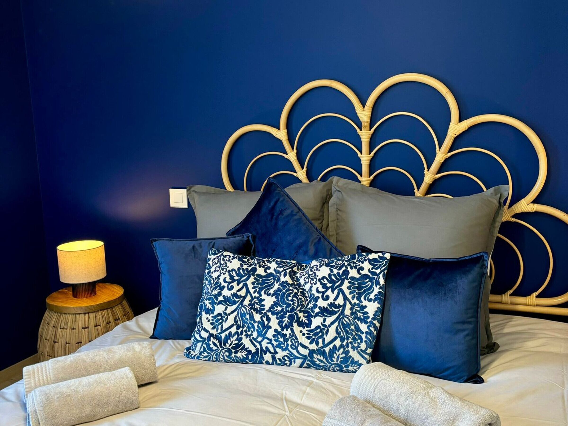 A bed with a blue and gold headboard, multiple pillows, a bedside lamp, and a wooden nightstand.