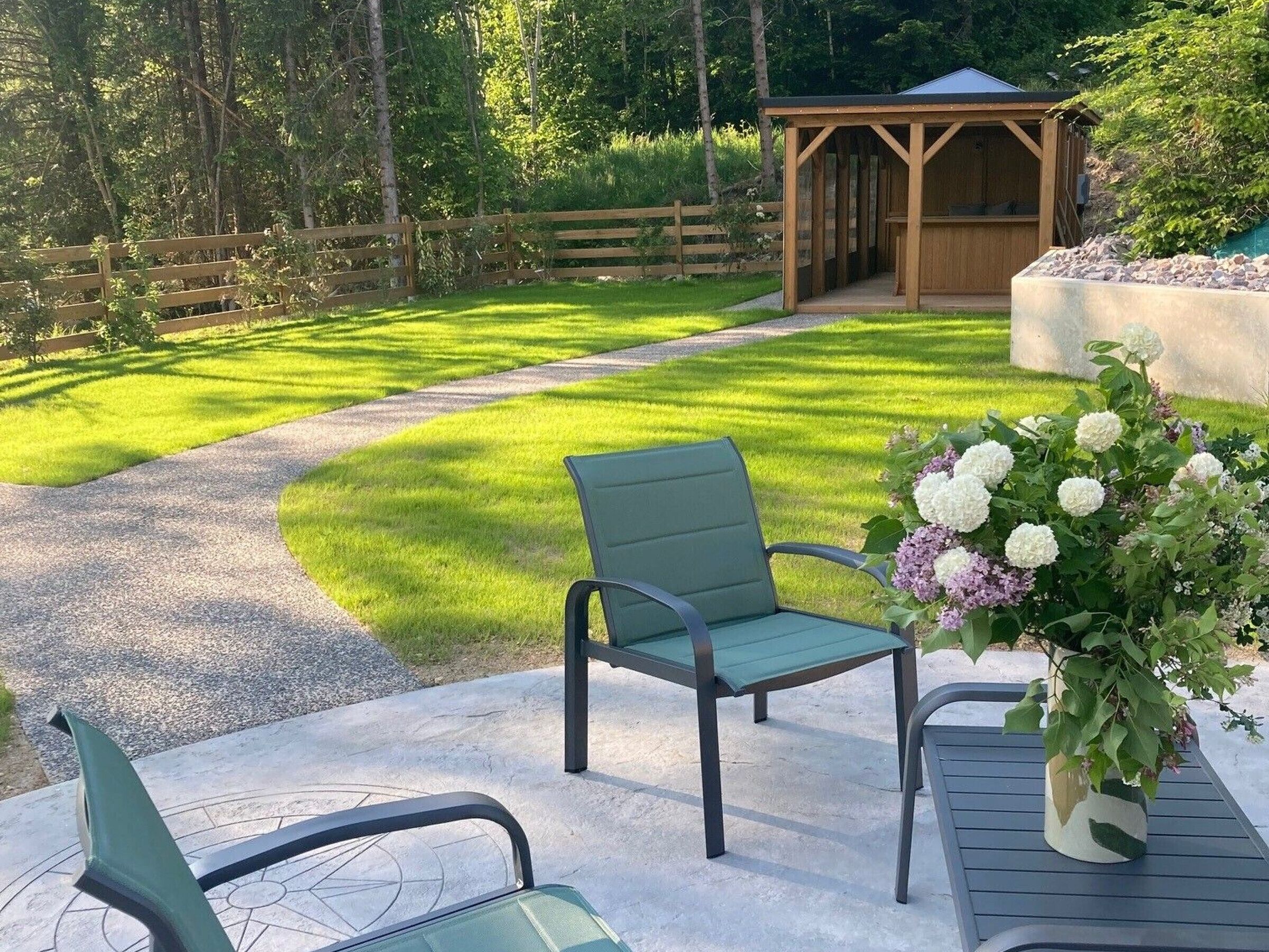 A patio with two green chairs and a table, a concrete path, a wooden fence, and a wooden structure in the background.