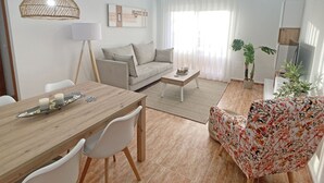 Una sala de estar moderna con sofá, mesa de centro, mesa de comedor y sillas. Se observa un ventanal con cortinas translúcidas, una lámpara colgante y una planta en maceta.