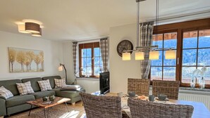A cozy living room with a sofa, coffee table, and wicker chairs. There is a television, a clock, and a window with a view of the mountains.