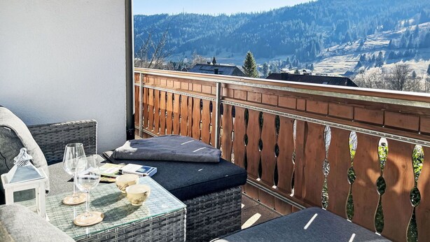 A balcony with wicker furniture, a table with wine glasses and bowls, and a view of a mountainous landscape.