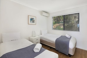 Quarto de hotel com duas camas, uma mesa de cabeceira, um quadro emoldurado, aparelho de ar condicionado e janela com persianas.