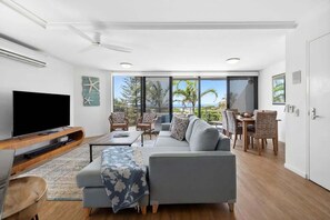A modern living room with a flat-screen TV, a sofa, a dining area with chairs, and large windows offering a view of palm trees and the ocean.