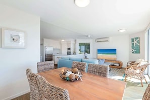 Un salon moderne avec une table à manger, des chaises en osier, une cuisine en arrière-plan et une grande fenêtre offrant une belle vue.