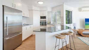 A modern kitchen with a large refrigerator, oven, and microwave. There is a bar stool area and a window with blinds. - Beautiful Belmore Retreat Easy Walk to Beach (Noosa)