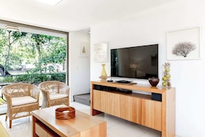 Un salon moderne dotĂ© dâun grand tĂ©lĂ©viseur Ă Ă©cran plat, de fauteuils en osier, dâune table basse en bois et dâune porte en verre donnant sur un jardin.