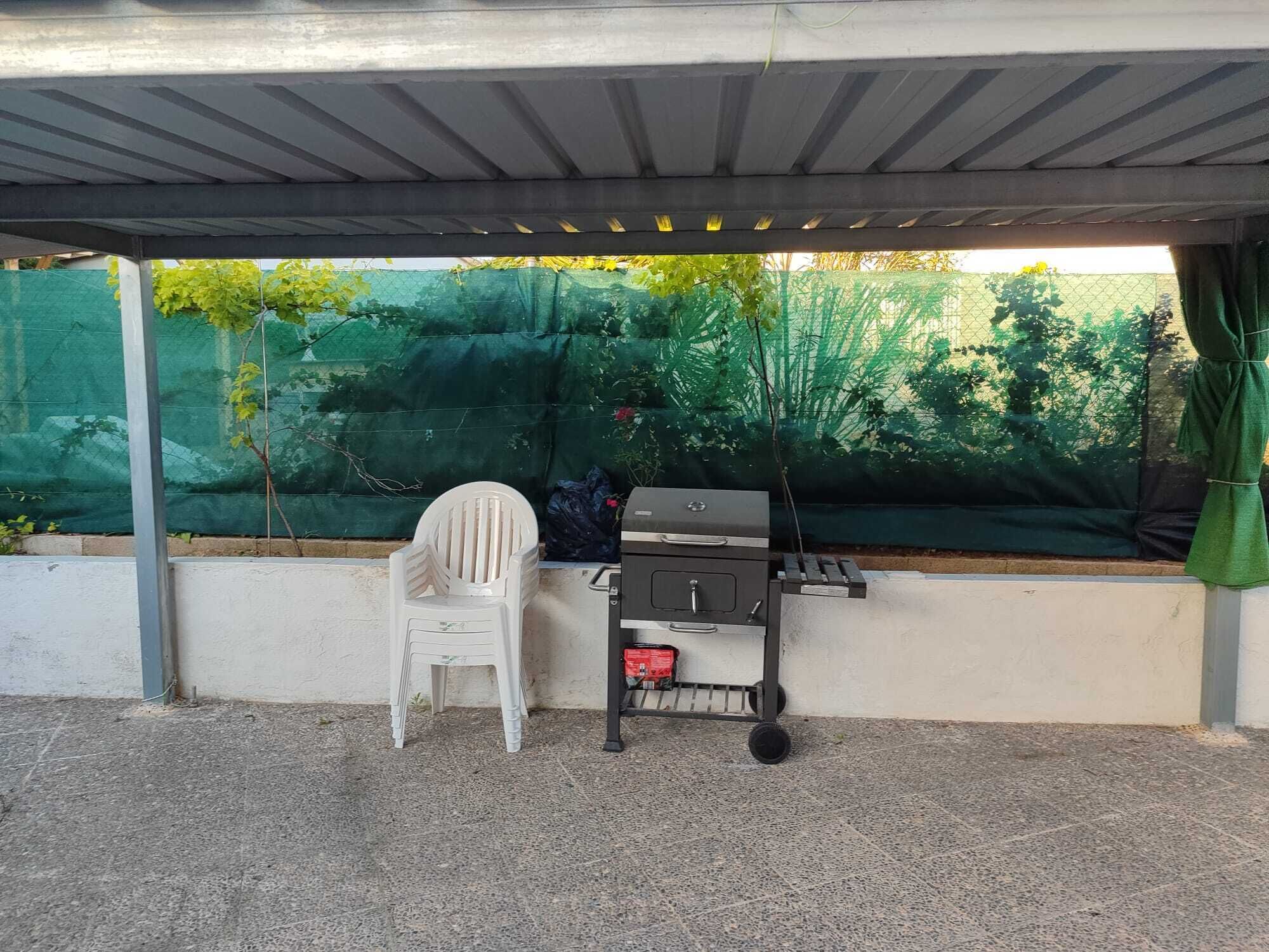 Ein überdachter Außenbereich mit Grill, einem weißen Plastikstuhl und einem grünen Netzhintergrund.