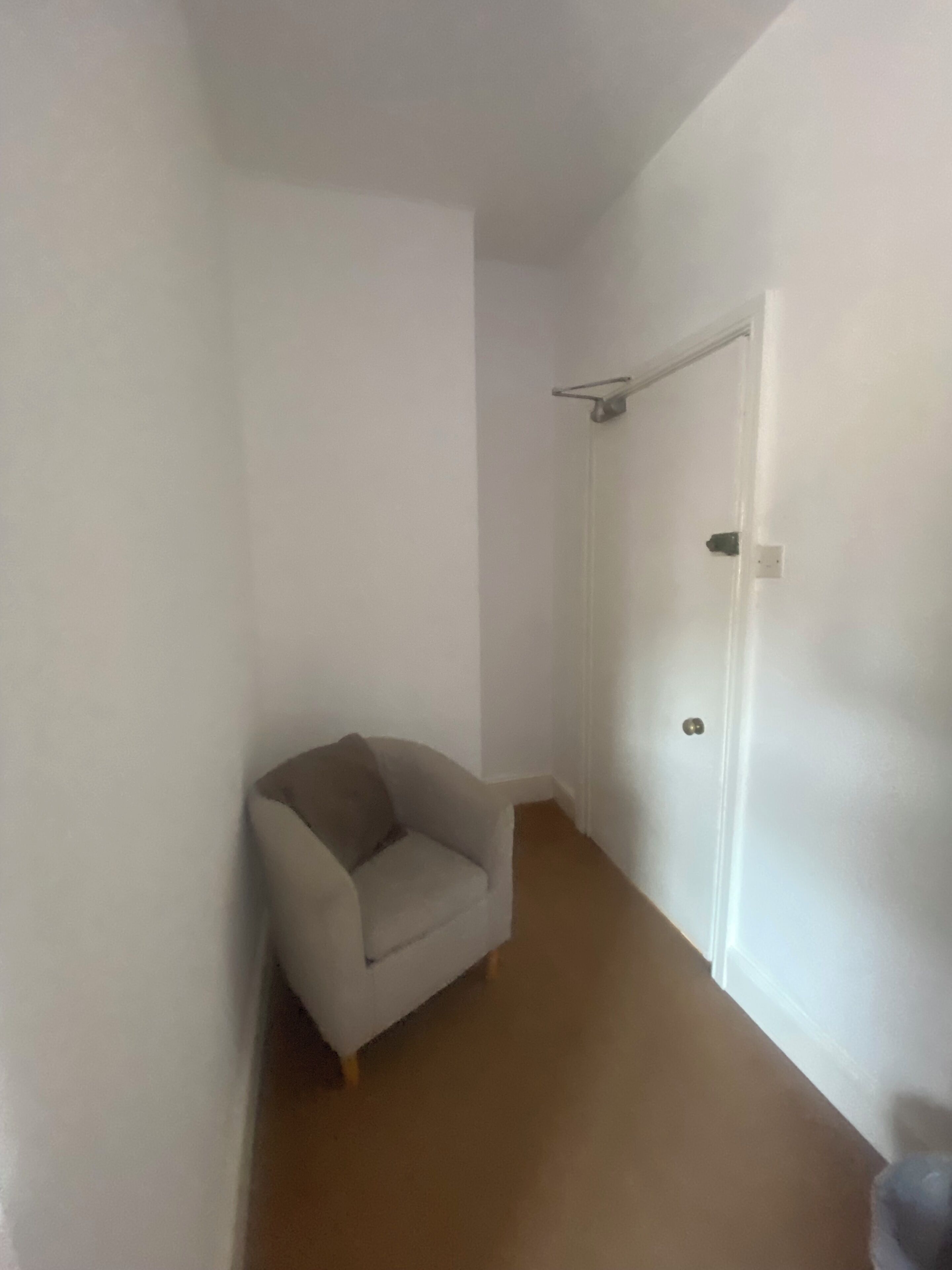 A narrow hallway with a white door at the end, a beige armchair, and a coat rack.