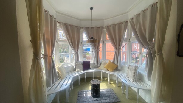A cozy room with a curved white bench, a small round table, and a view of the outside through large windows with curtains. - Stokesay inn and b and b  (Craven Arms)