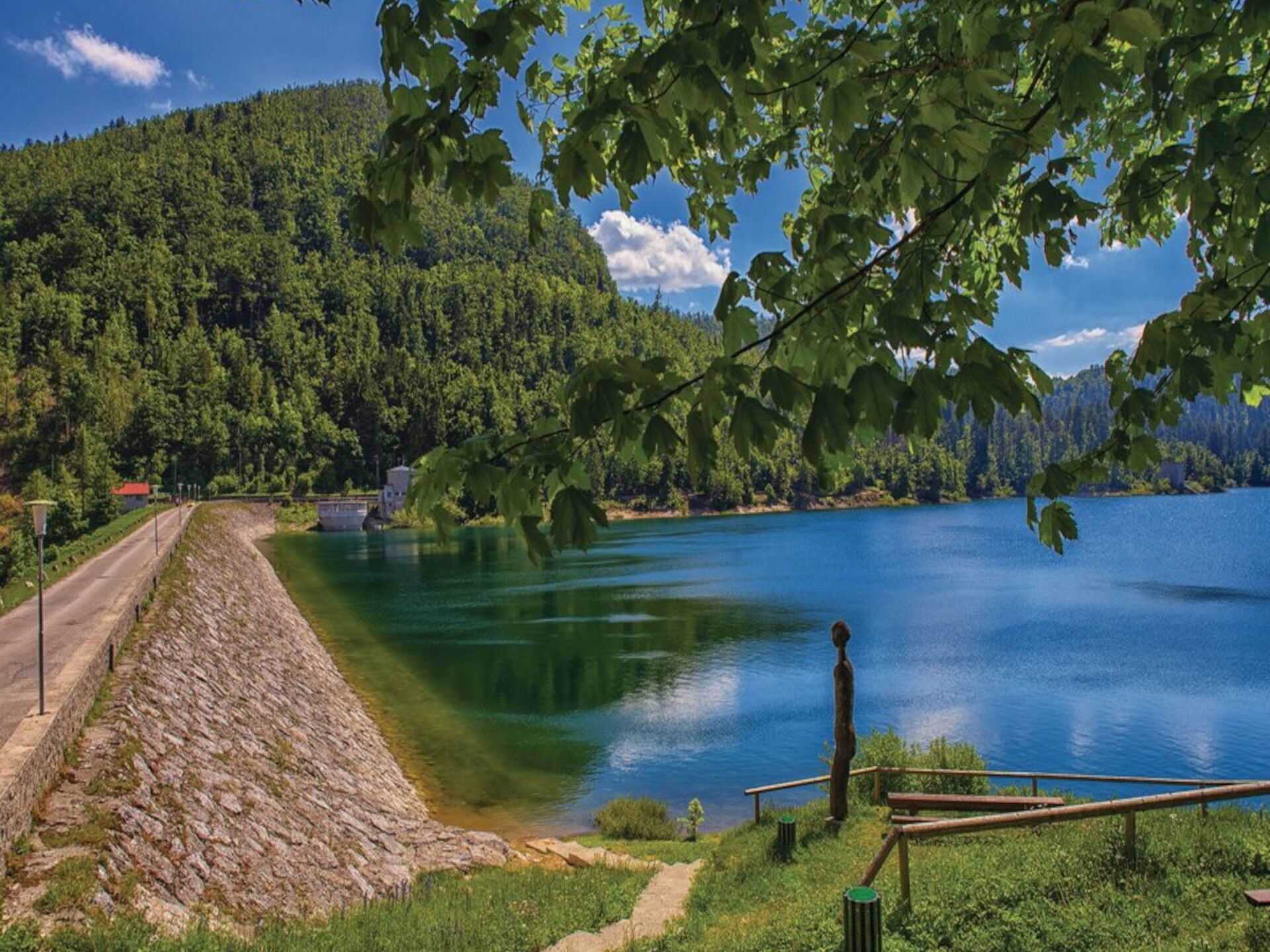 A serene lakeside view with a stone embankment, a wooden walkway, and a distant building.