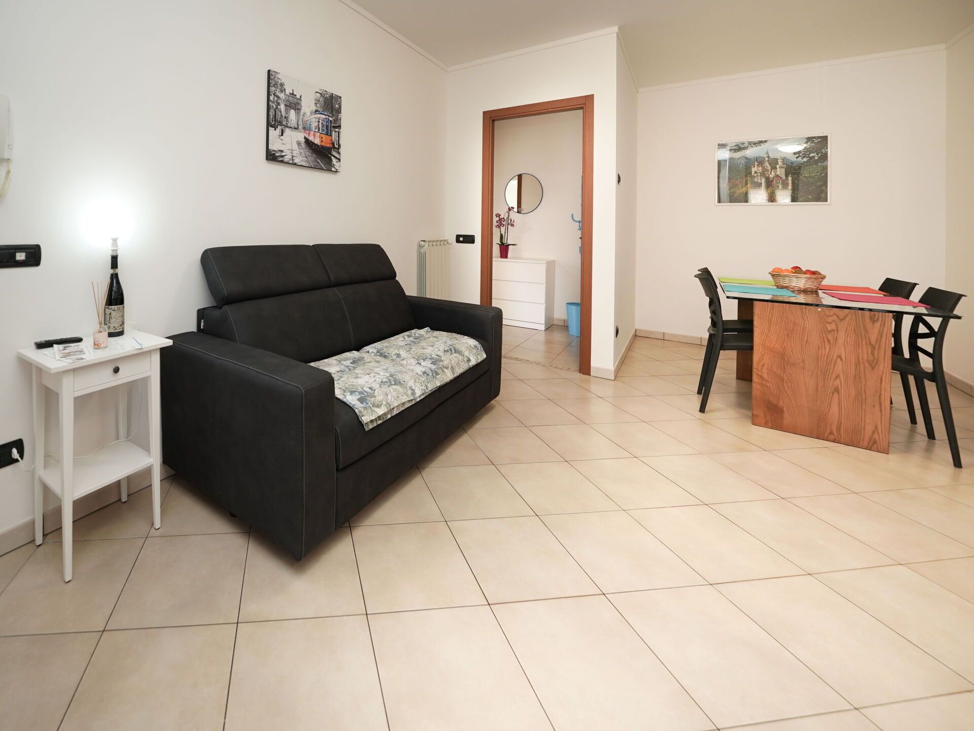 A living room with a black sofa, a small white side table, a wooden dining table with chairs, and two wall-mounted pictures.