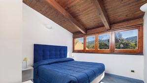 A bedroom with a large bed, wooden ceiling beams, a window with a view, and a bedside table with a lamp.