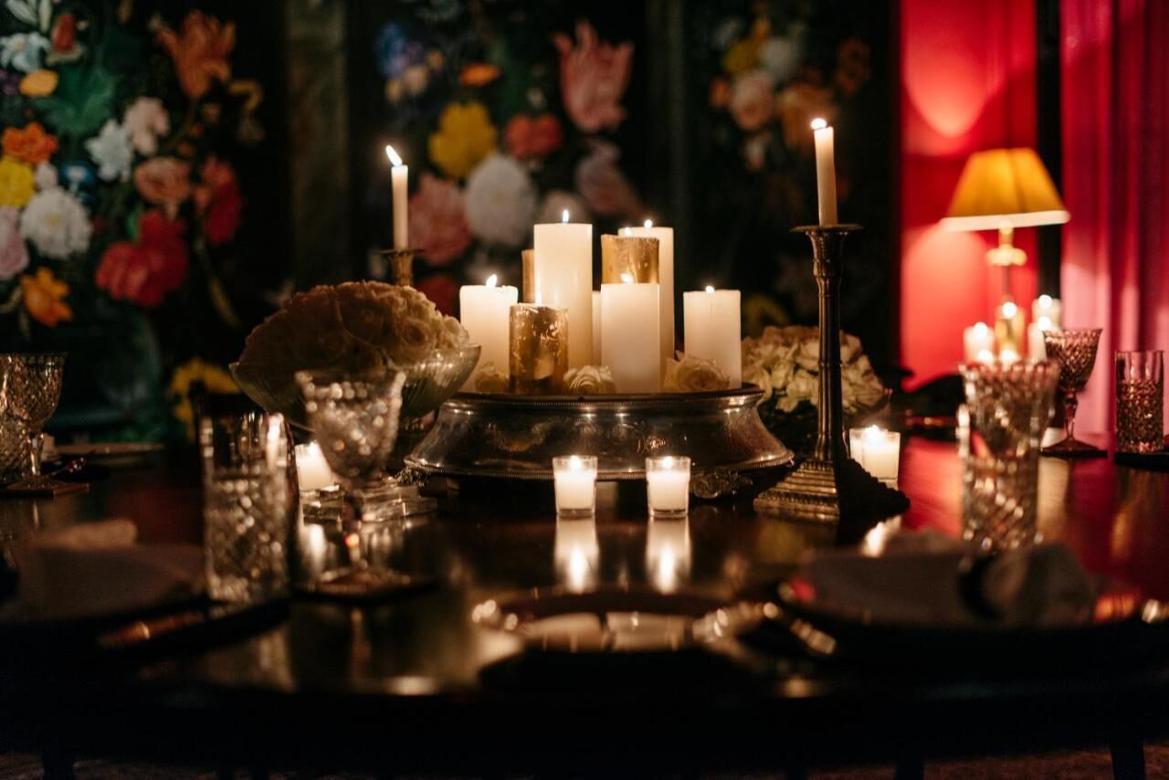 Una mesa dispuesta con velas, una bandeja de latón y cristalería, con un fondo floral y una lámpara al fondo.
