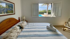 A bedroom with a bed, a window with curtains, a painting on the wall, and a view of a pool and palm trees outside.