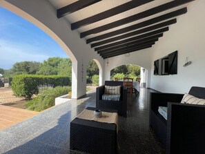 Une terrasse extérieure avec des meubles en osier noir, une table et un banc. On y voit des arches et, au fond, une vue sur la verdure.