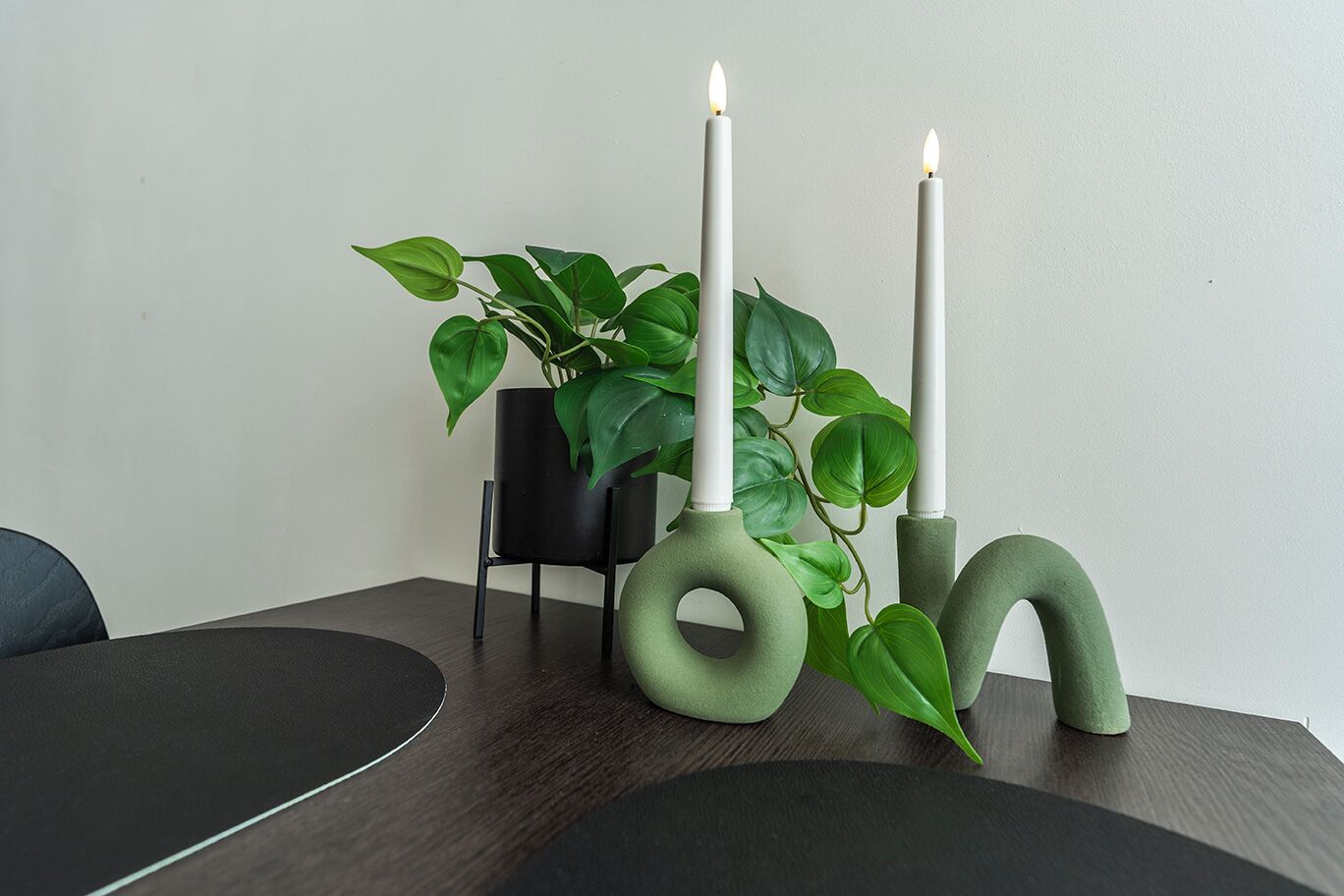Een minimalistisch ingerichte kamer met een donker houten tafel, twee groene kandelaars en een potplant.