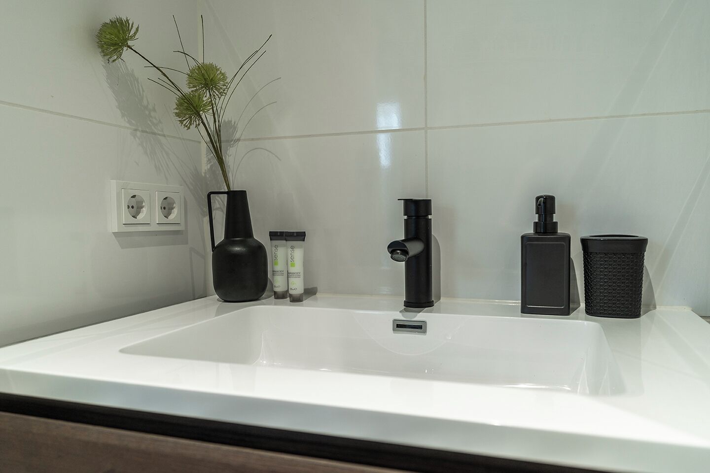 A bathroom with a white sink, a black faucet, a black vase with green flowers, and two bottles of shampoo.