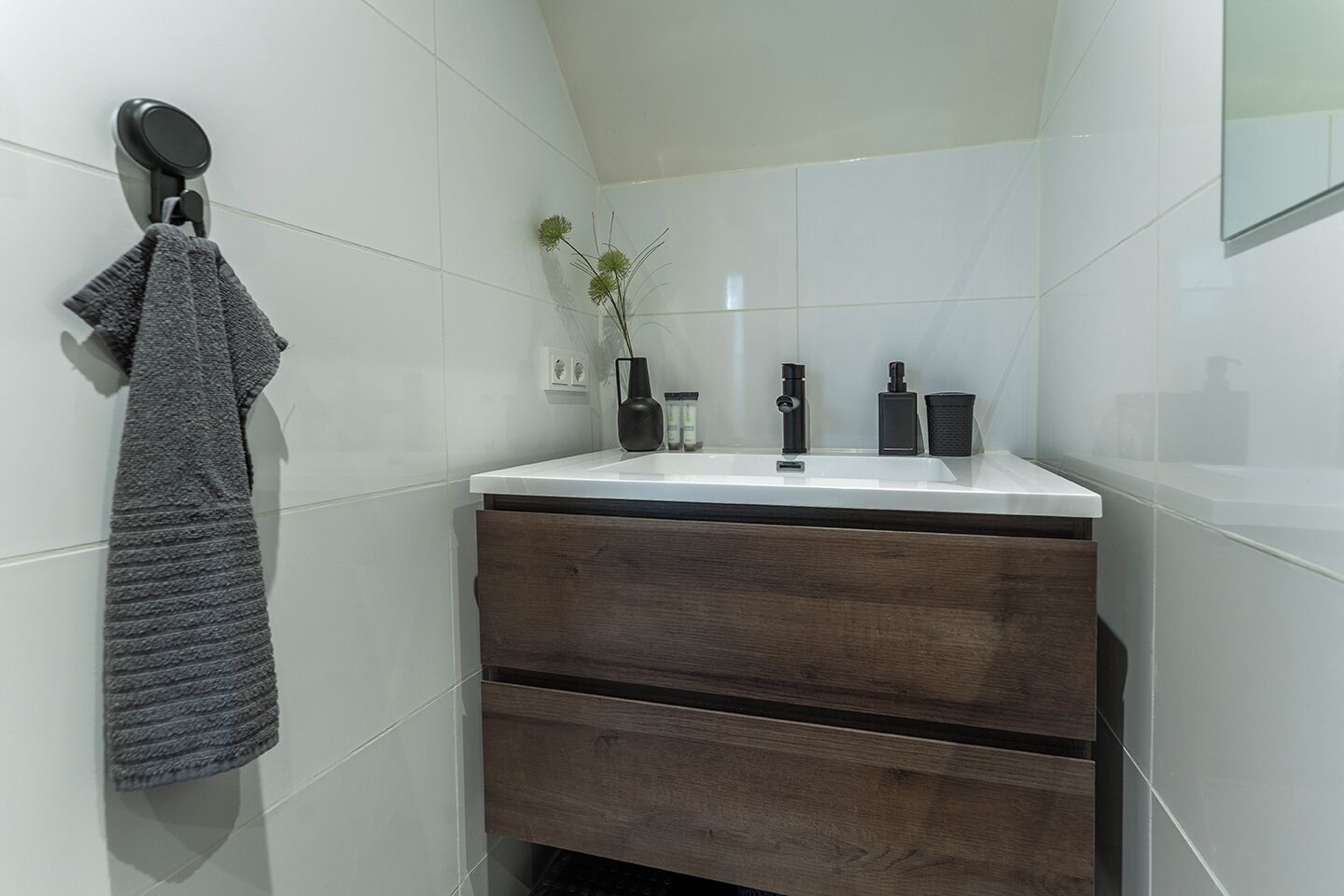 A modern bathroom with a white sink, dark wood drawers, a towel rack, a vase with flowers, and a mirror.