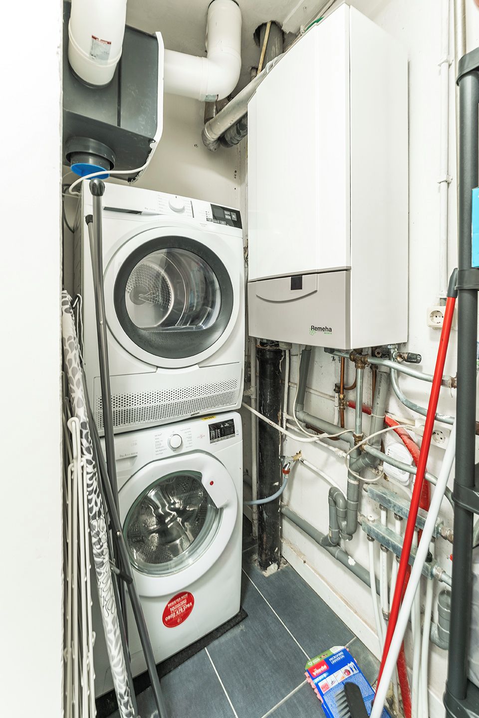 Een kleine wasruimte met twee gestapelde wasmachines, een boiler en zichtbare leidingen.