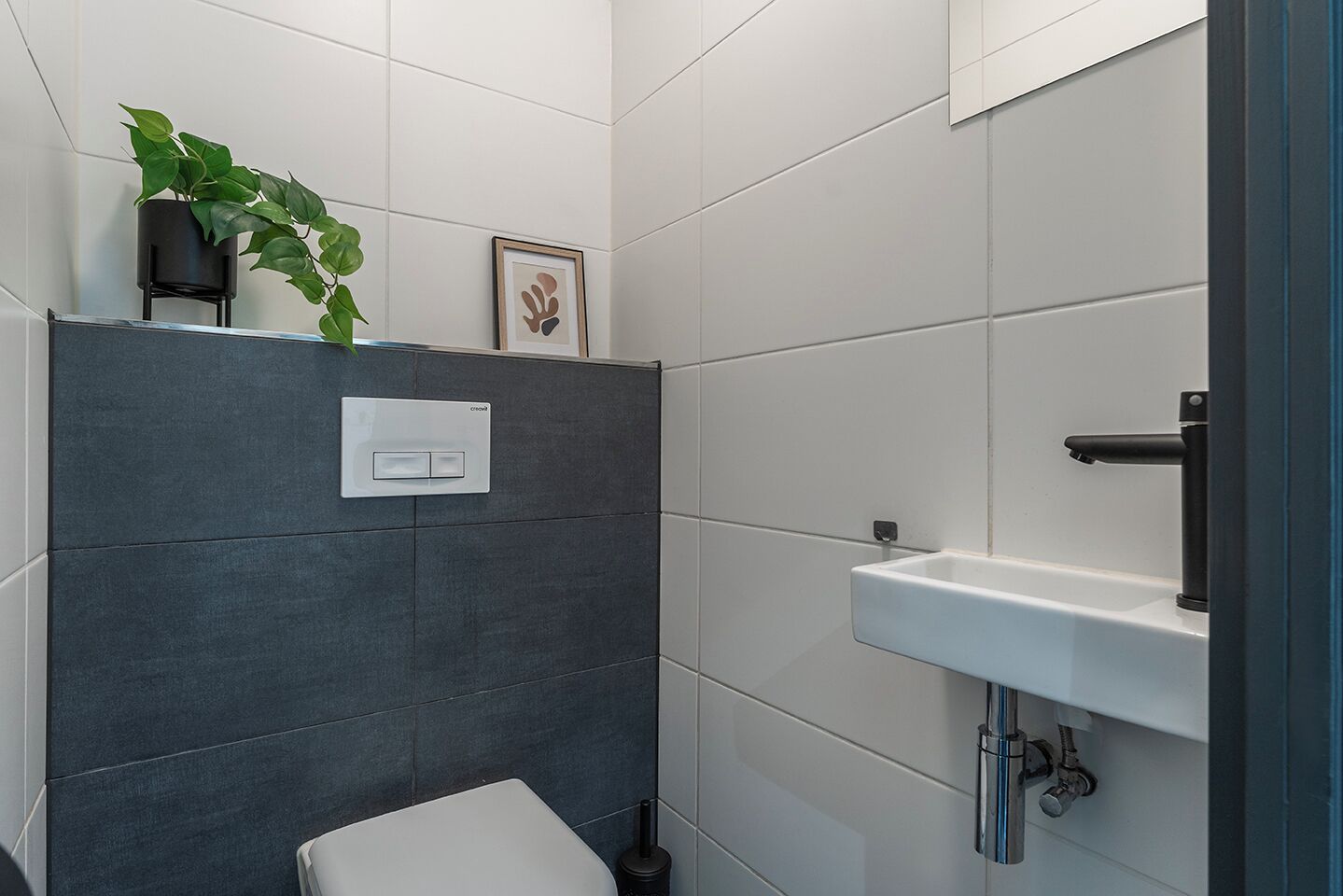 Ein modernes Badezimmer mit grauen Fliesen, einer weißen Toilette, einem Waschbecken mit chromfarbenem Wasserhahn, einer Topfpflanze und einem gerahmten Bild.