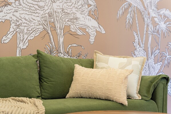 Un canapé vert avec deux coussins, placé contre un mur orné d’une fresque aux motifs tropicaux.