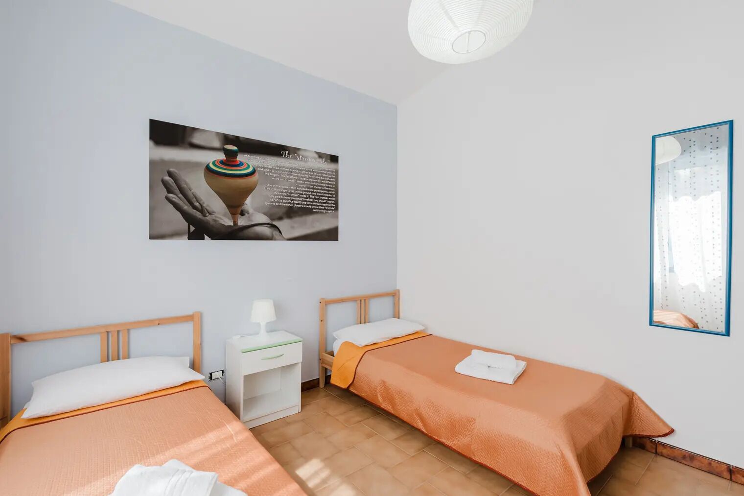 A hotel room with two single beds, a nightstand, a lamp, a framed picture on the wall, and a window with a view.
