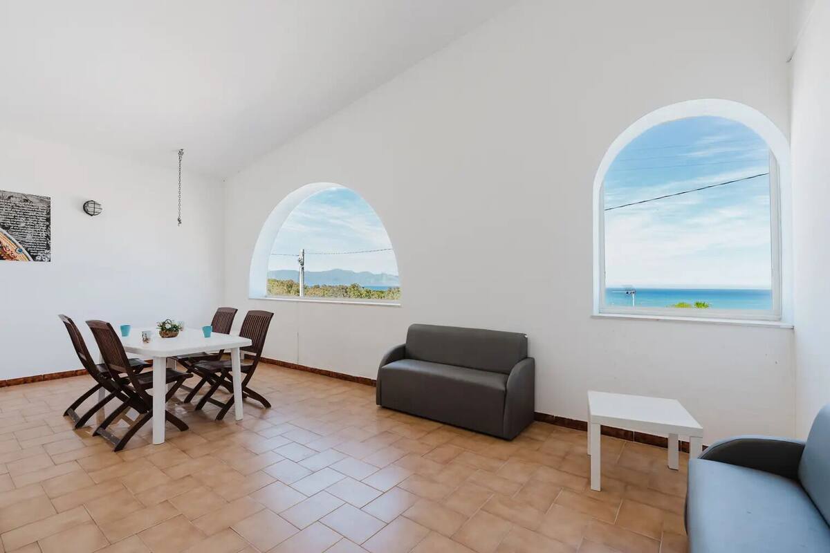 Une pièce spacieuse comprenant une table à manger et des chaises, un canapé gris et deux fenêtres en arc d’où l’on peut admirer la mer.