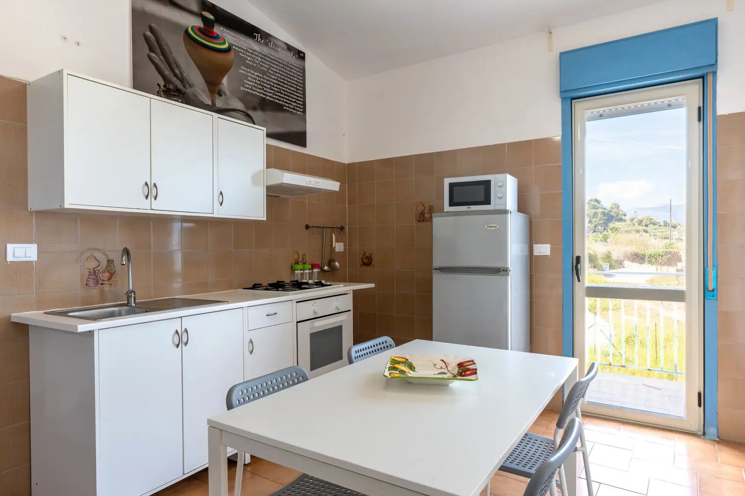 A modern kitchen with white cabinets, a gas stove, a microwave, and a refrigerator. There's a dining table with chairs and a view of the outdoors through a sliding glass door.