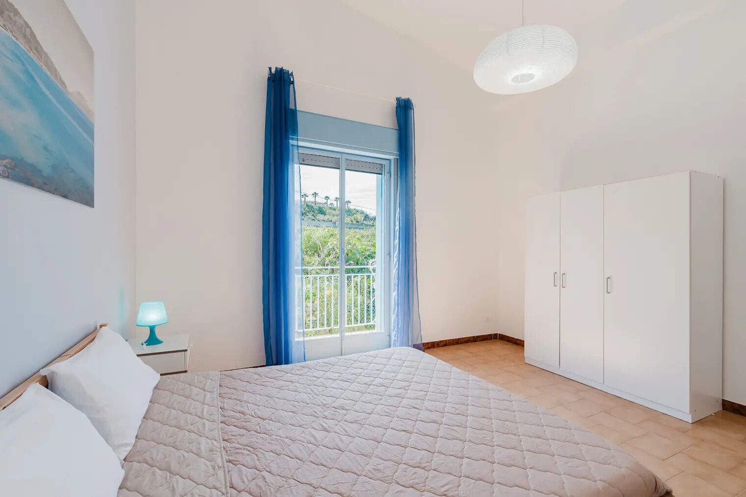 A bedroom with a bed, a nightstand with a lamp, a wardrobe, and a balcony with blue curtains.