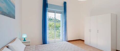 A bedroom with a bed, a nightstand with a lamp, a wardrobe, and a balcony with blue curtains.