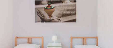 A hotel room with two beds, a bedside table with a lamp, and a wall-mounted picture of a hand holding a pottery piece.
