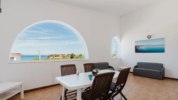 Una habitación con una mesa de comedor, sillas, un sofá y un amplio ventanal de arco con vistas al mar.