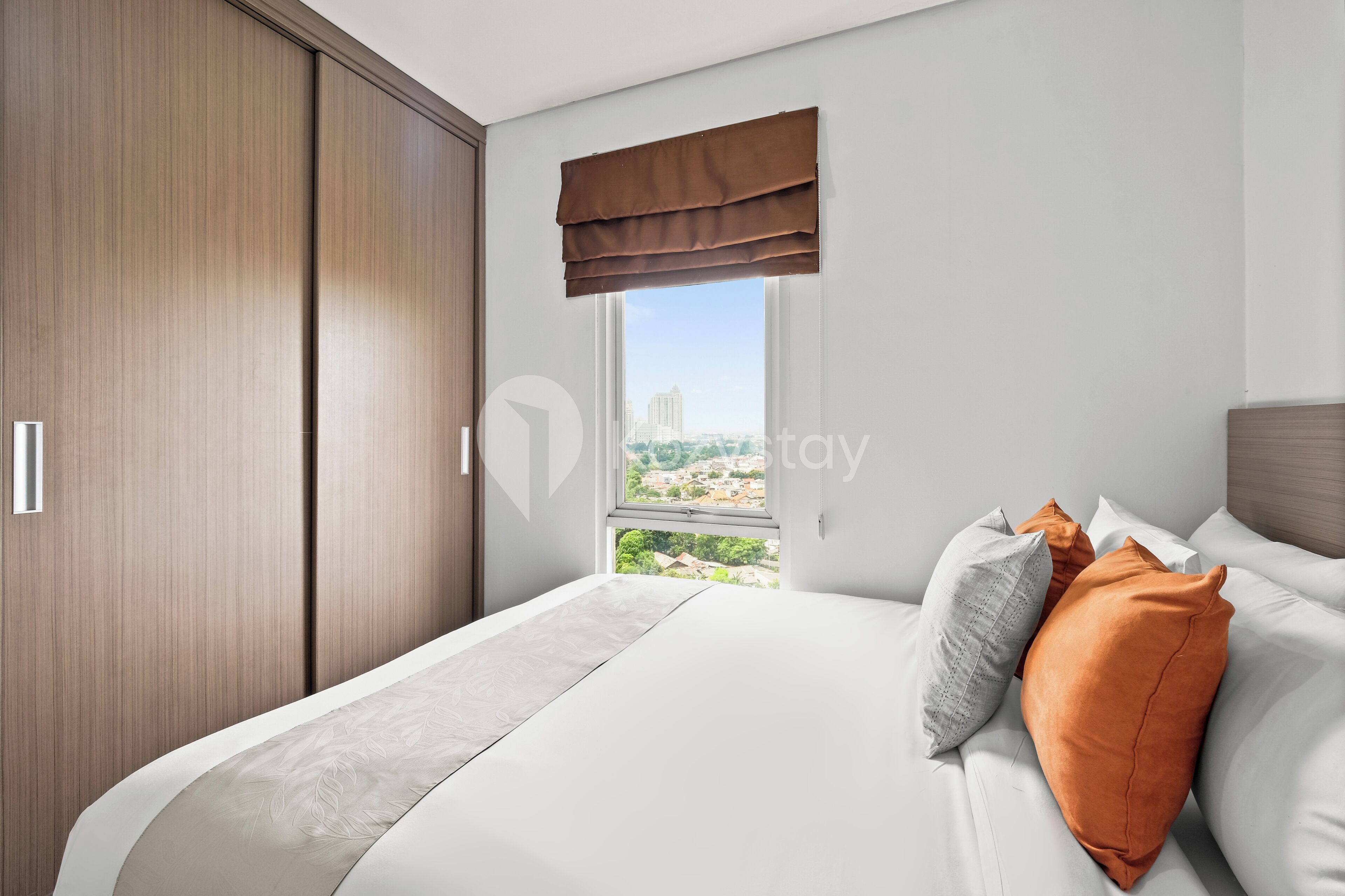 Un dormitorio con un ventanal grande, una cama con ropa de cama blanca y almohadas naranjas, y un armario de madera.