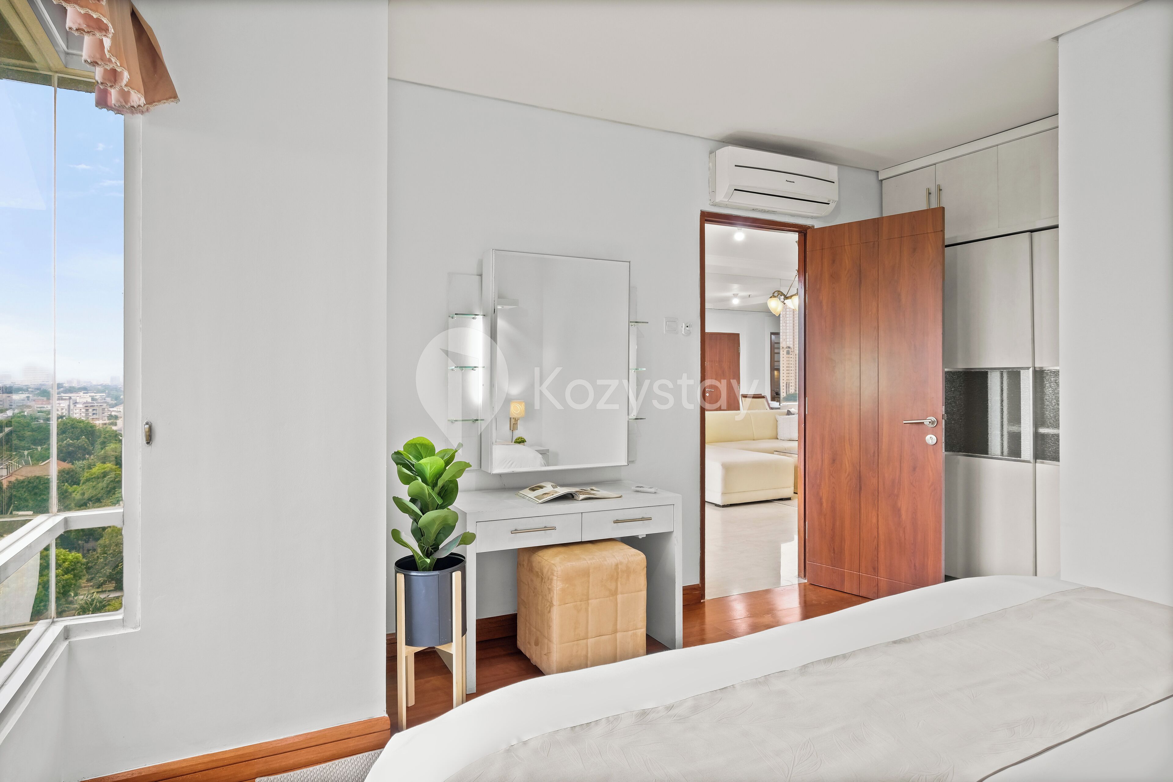 A modern bedroom with a bed, a dresser, a vanity with a mirror, a potted plant, and a view of the city.