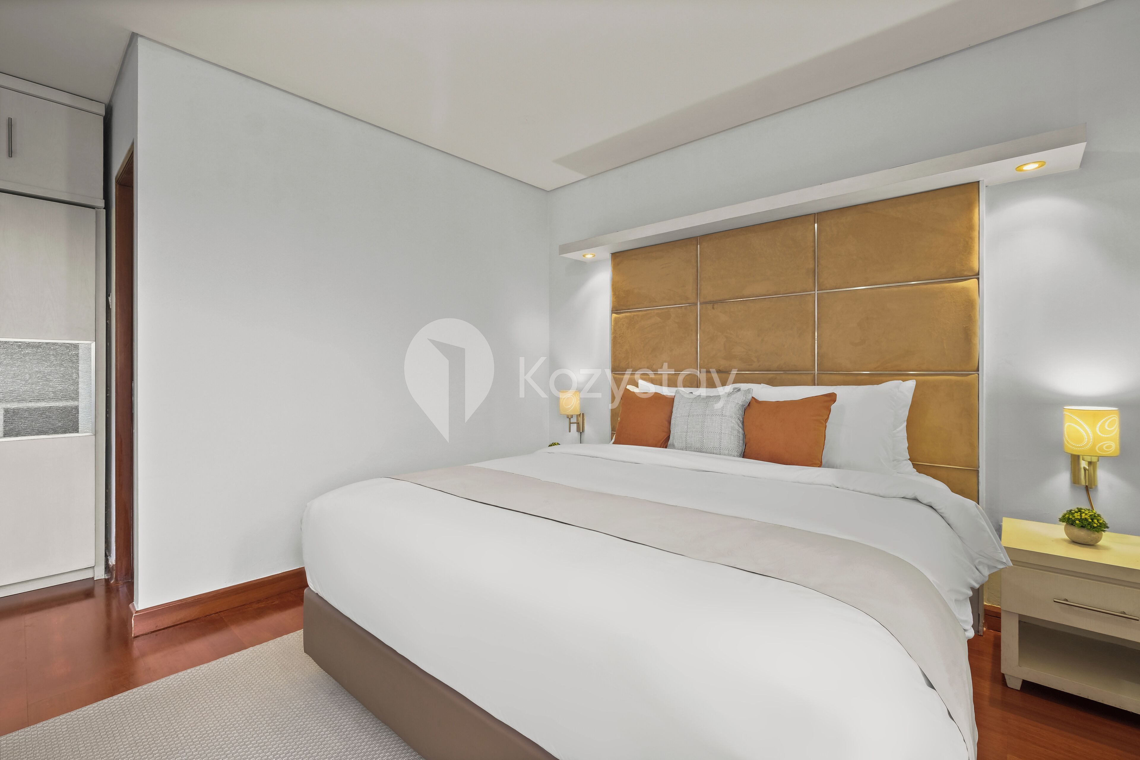 A modern hotel room with a large bed, wooden headboard, bedside lamps, and a small table with a plant.